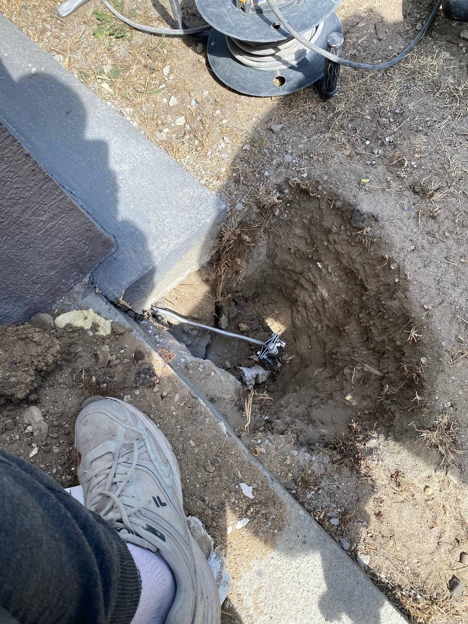Uziemienie w wykopie przy betonowym obrzeżu. Widoczny kabel na szpuli i fragment buta. Instalacja elektryczna w ziemi.