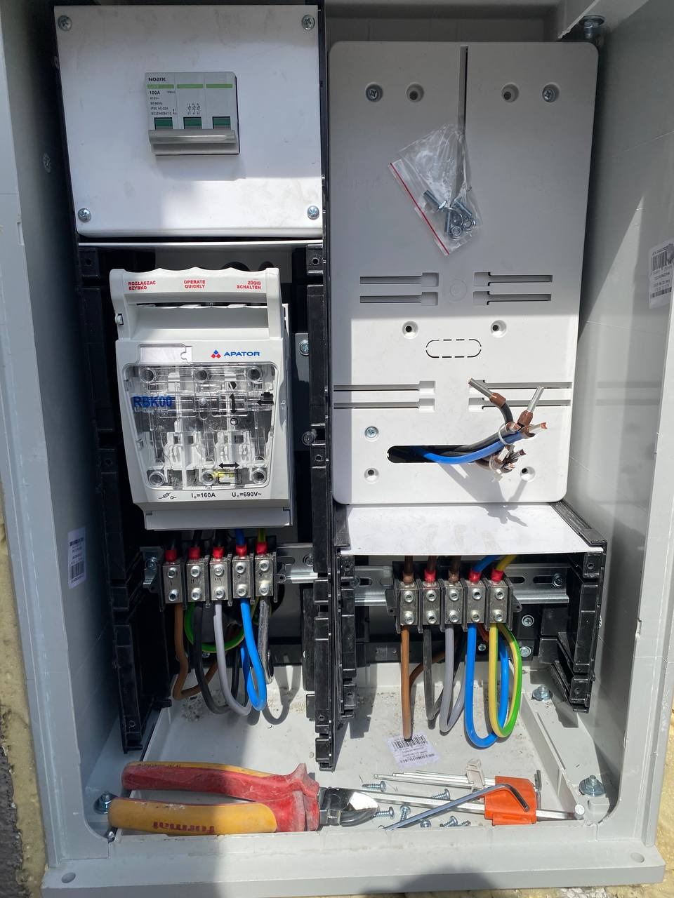Otwarta, szara skrzynka elektryczna z zamontowanymi bezpiecznikami, przewodami i narzędziami (kombinerki, klucz imbusowy). Widoczne złącza i okablowanie w kolorach niebieskim i brązowym.