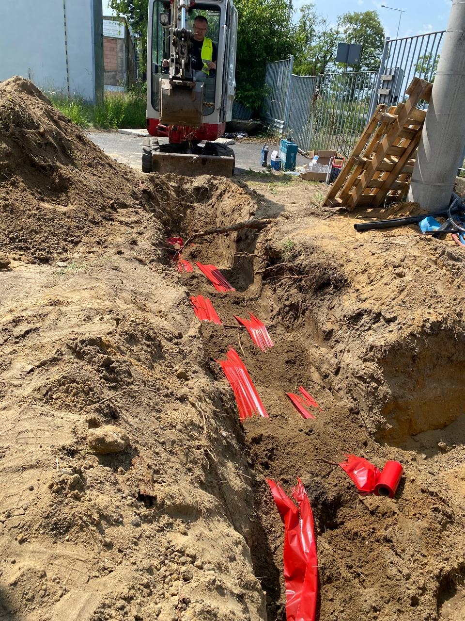 Wykop z czerwonymi taśmami ostrzegawczymi, prawdopodobnie zabezpieczającymi kable elektryczne, z minikoparką w tle i betonowym słupem po prawej.