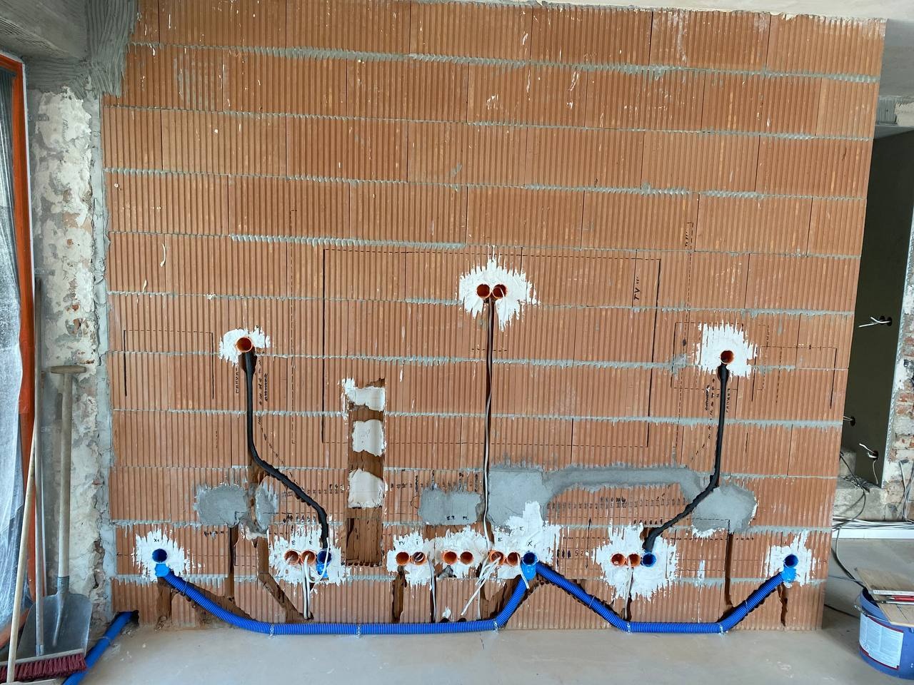 Ceglana ściana z instalacją elektryczną: kable w peszlach, puszki zalane gipsem. Widoczne narzędzia malarskie i murarskie. Etap prac przed tynkowaniem.