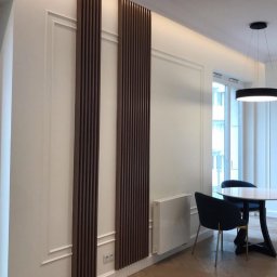 Adam Sadulaev - Nowoczesne panele dekoracyjne z drewna na ścianie w jasnym wnętrzu. Widoczny stół z krzesłami i oświetlenie sufitowe.