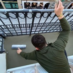 Adam Sadulaev - Prace tynkarskie na balkonie w Warszawie: mężczyzna w zielonej bluzie wyrównuje ścianę szpachelką, widok z góry na balkon z metalową balustradą.