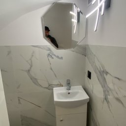 Adam Sadulaev - Nowoczesna łazienka z umywalką na szafce, marmurowe płytki, lustro w kształcie ośmiokąta i designerska lampa LED. Widoczne odbicie osoby w lustrze.