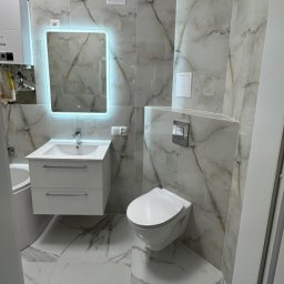 Adam Sadulaev - Elegancka łazienka w bieli i szarości z marmurkowym wzorem płytek, podwieszana toaleta, szafka z umywalką i lustro z podświetleniem LED.