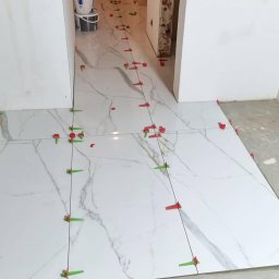 Adam Sadulaev - Białe płytki imitujące marmur, układane w korytarzu. System poziomowania płytek z czerwonymi i zielonymi klipsami. Widoczne linie fug. Etap prac glazurniczych.