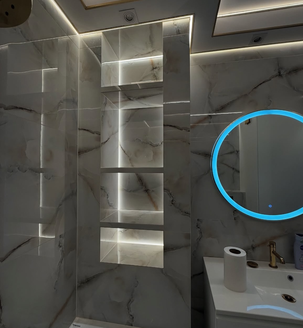 Wykończona łazienka z marmurowymi płytkami, podświetlanymi półkami i okrągłym lustrem z niebieską obwódką. Nowoczesny design z dbałością o detale i oświetlenie.