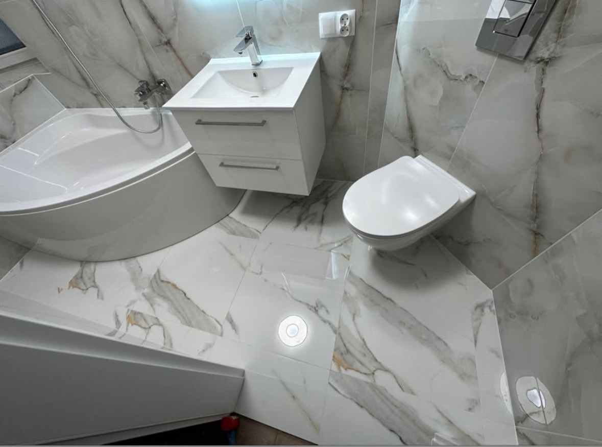 Biała, nowoczesna łazienka z marmurowymi płytkami: wanna, umywalka z szafką i podwieszana toaleta. Widok z góry, oświetlenie punktowe w podłodze.