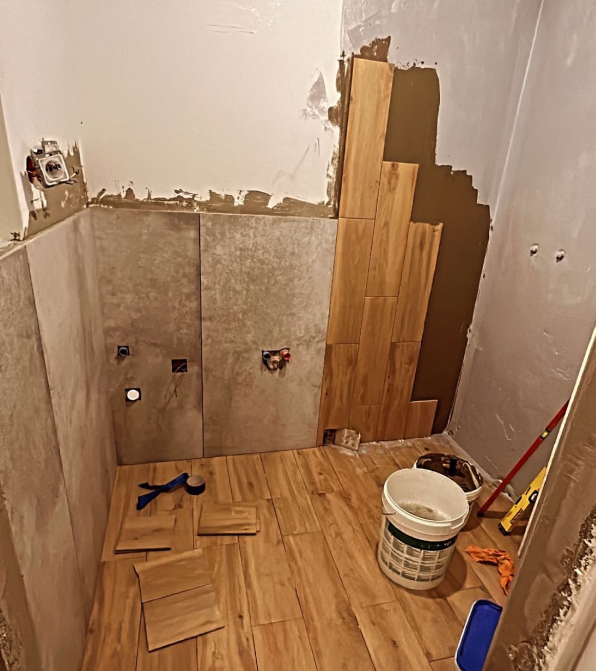 Układanie drewnopodobnych płytek w łazience. Częściowo wykończone ściany i podłoga. Widoczne narzędzia i wiadro z klejem. W tle niezakończone prace.