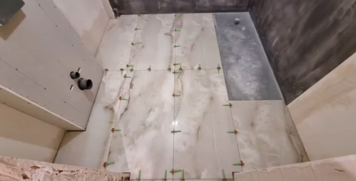 Układanie płytek podłogowych w łazience, z widocznymi krzyżykami dystansowymi. Częściowo wykończona ściana z płyt karton-gips i fragment wykończonej ściany w szarości.