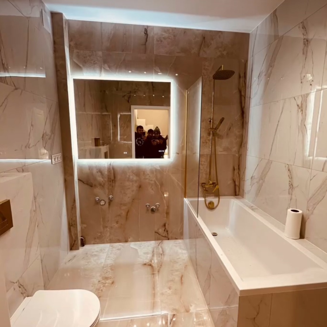 Wykończona łazienka w marmurze z podświetlanym lustrem, złotą armaturą i widoczną toaletą. W lustrze odbija się grupa osób. Nowoczesny i elegancki design.