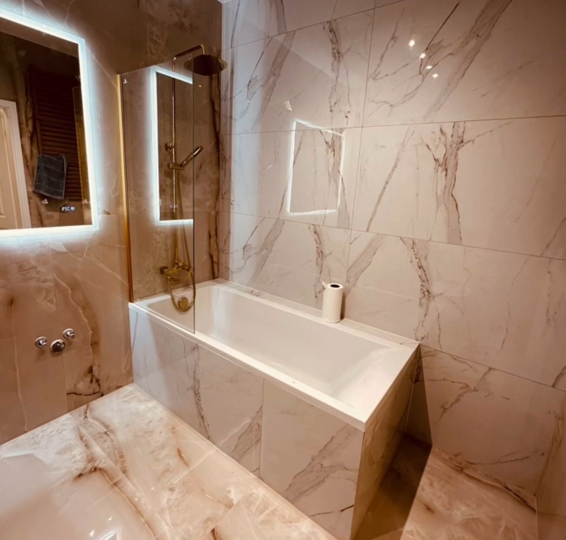 Wykończona łazienka z wanną obudowaną marmurowymi płytkami, złota armatura, podświetlane lustro i wnęka. Nowoczesny design z akcentem luksusu.