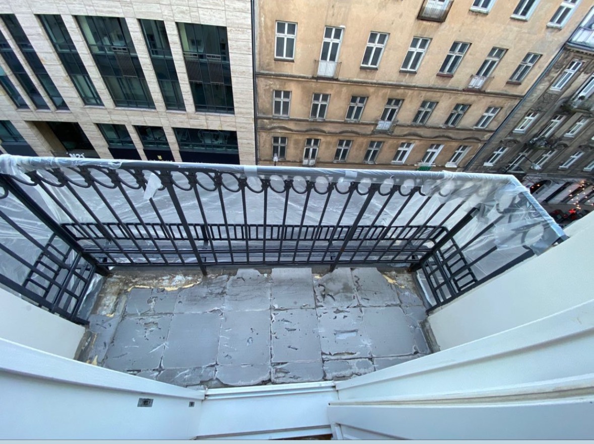 Balkon w trakcie remontu z odsłoniętą, szarą posadzką. Metalowa balustrada zabezpieczona folią. W tle budynki o zróżnicowanej architekturze.