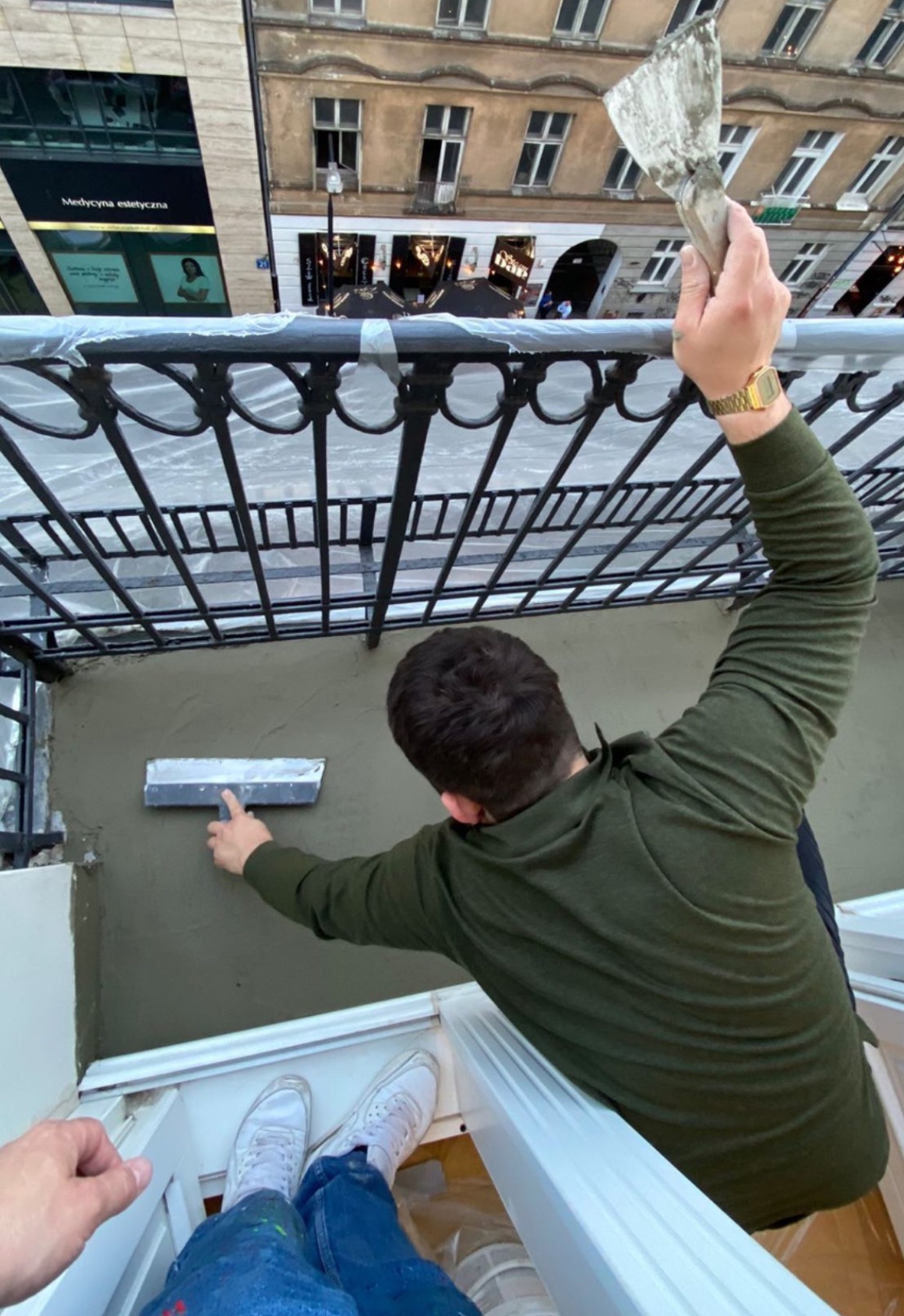 Prace tynkarskie na balkonie w Warszawie: mężczyzna w zielonej bluzie wyrównuje ścianę szpachelką, widok z góry na balkon z metalową balustradą.