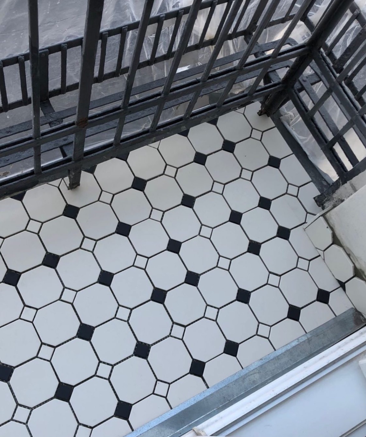 Ukończony remont balkonu: biało-czarna mozaika podłogowa w geometryczny wzór, widok z góry, metalowa balustrada w tle, świeżo położone płytki.