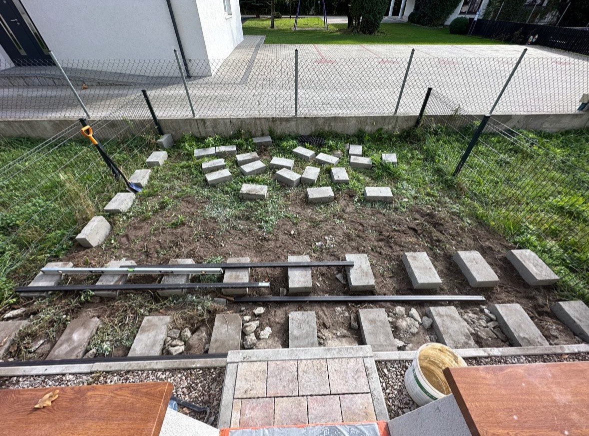 Układanie ścieżki z kostki brukowej w ogrodzie. Widoczne bloczki, poziomica i narzędzia. Etap przygotowawczy, częściowo ułożona kostka, ziemia i trawa w tle.