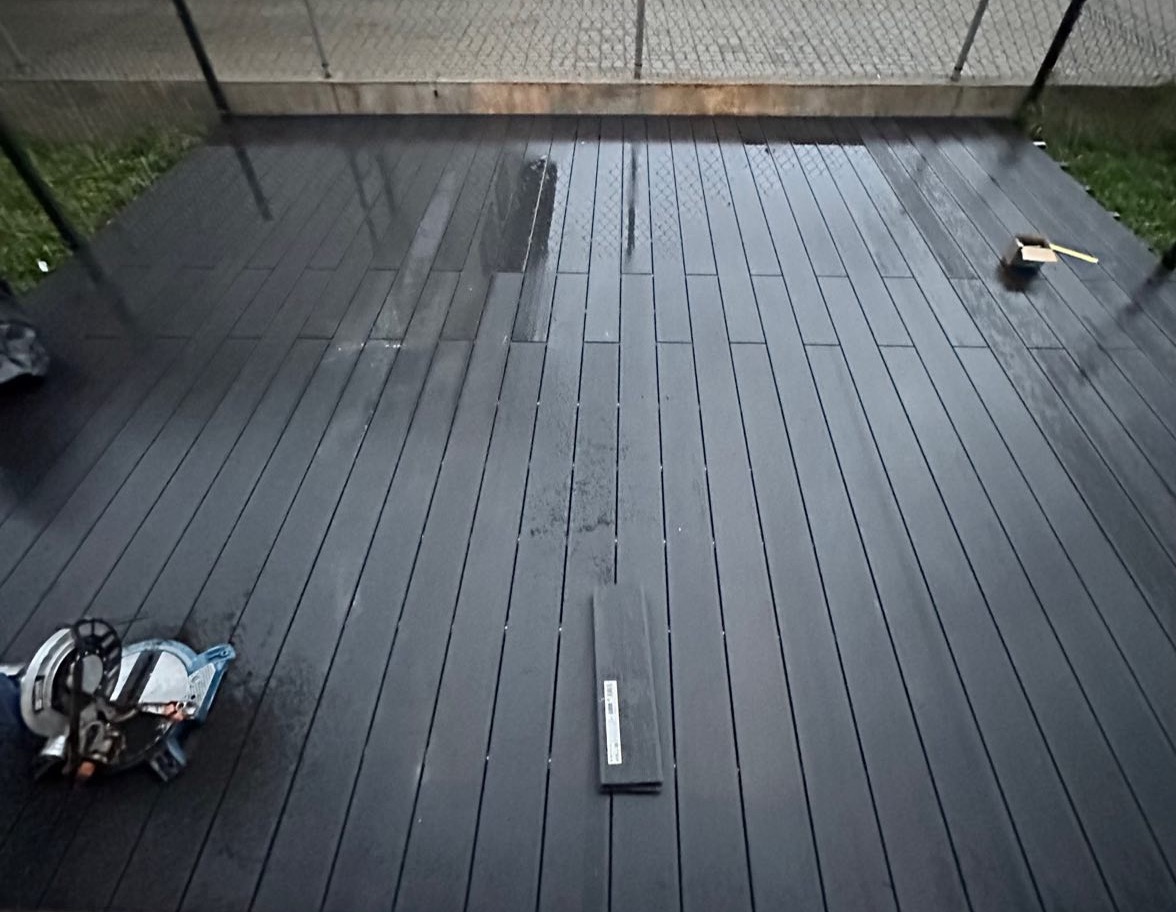 Montaż desek kompozytowych na balkonie, widoczna piła do cięcia, deski w trakcie układania. Ciemna kolorystyka, widok z góry na częściowo wykończoną podłogę.