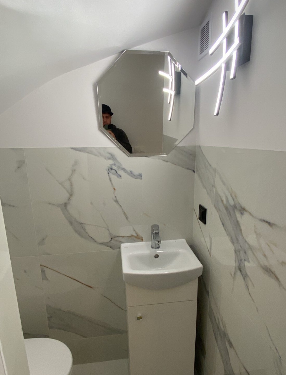 Nowoczesna łazienka z umywalką na szafce, marmurowe płytki, lustro w kształcie ośmiokąta i designerska lampa LED. Widoczne odbicie osoby w lustrze.