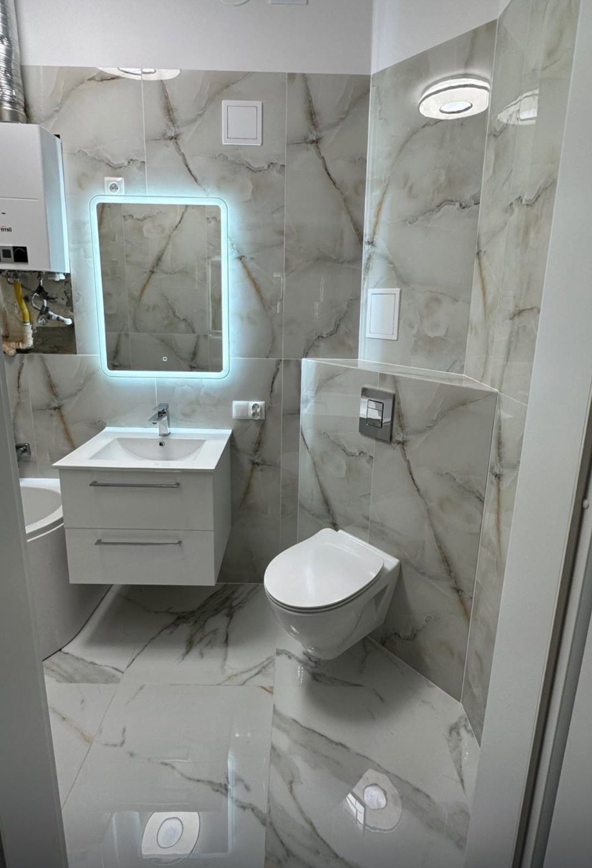 Elegancka łazienka w bieli i szarości z marmurkowym wzorem płytek, podwieszana toaleta, szafka z umywalką i lustro z podświetleniem LED.