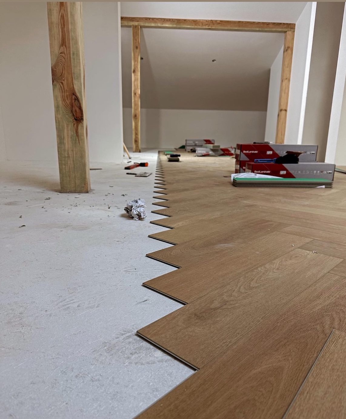 Układanie parkietu w jodełkę na poddaszu, widoczne deski i narzędzia. Częściowo wykończona podłoga, surowy beton, drewniane podpory konstrukcyjne.