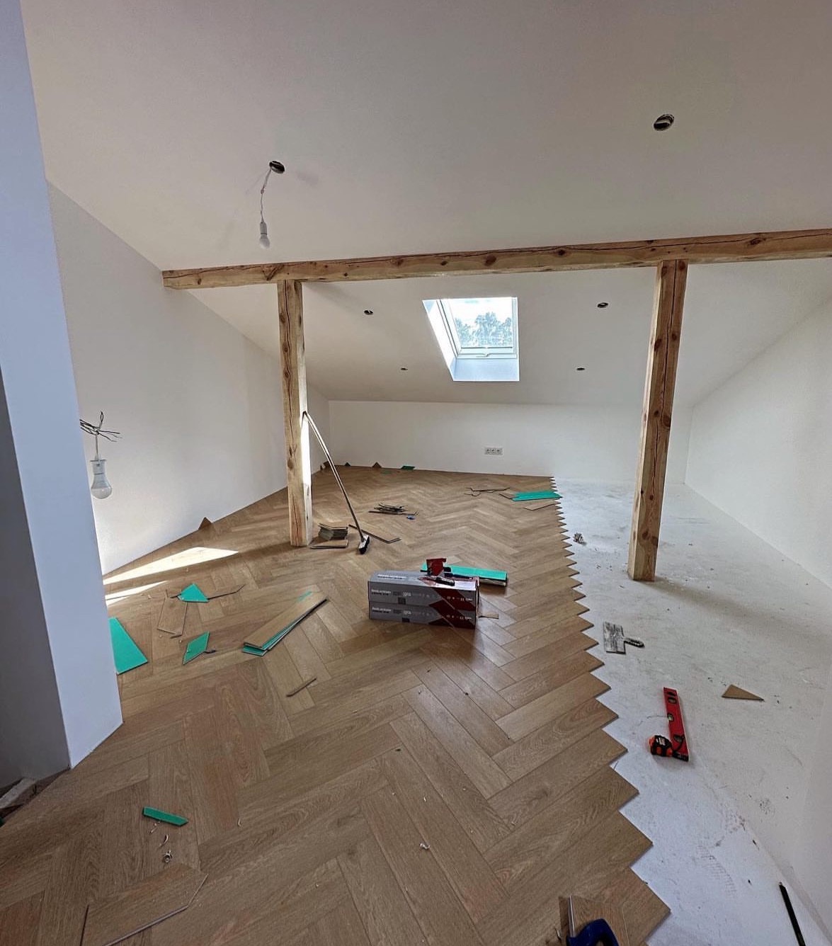 Układanie parkietu w jodełkę na poddaszu z drewnianymi belkami. Częściowo ułożona podłoga, widoczne narzędzia i materiały. Okno dachowe w tle.