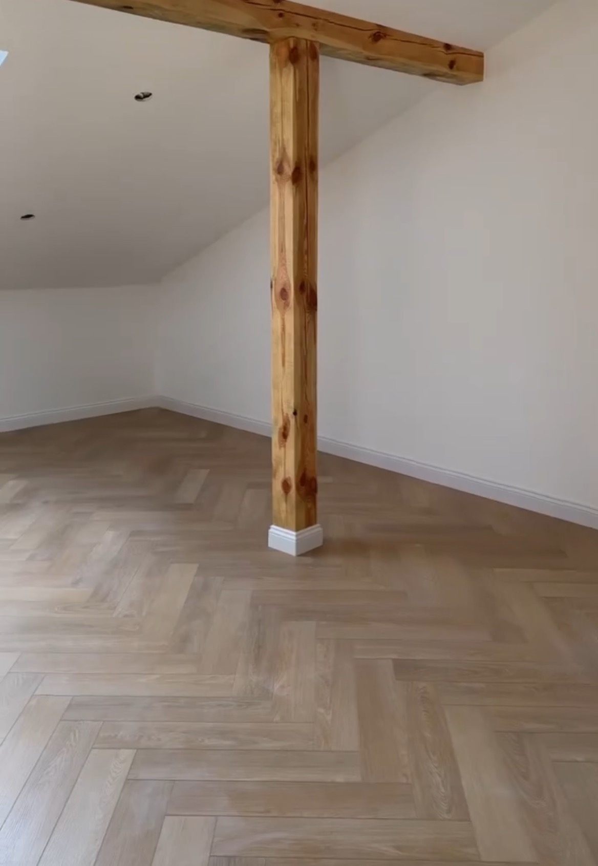 Jasny parkiet ułożony w jodełkę francuską w pokoju z drewnianą kolumną. Minimalistyczne wykończenie wnętrza poddasza z widocznym drewnem konstrukcyjnym.