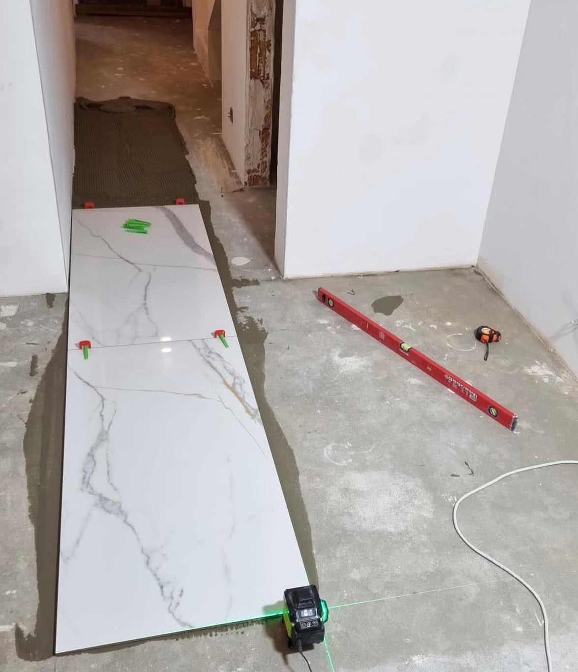 Układanie płytek imitujących marmur w korytarzu, poziomica laserowa, system poziomowania płytek. Widoczny klej do płytek i poziomica tradycyjna.