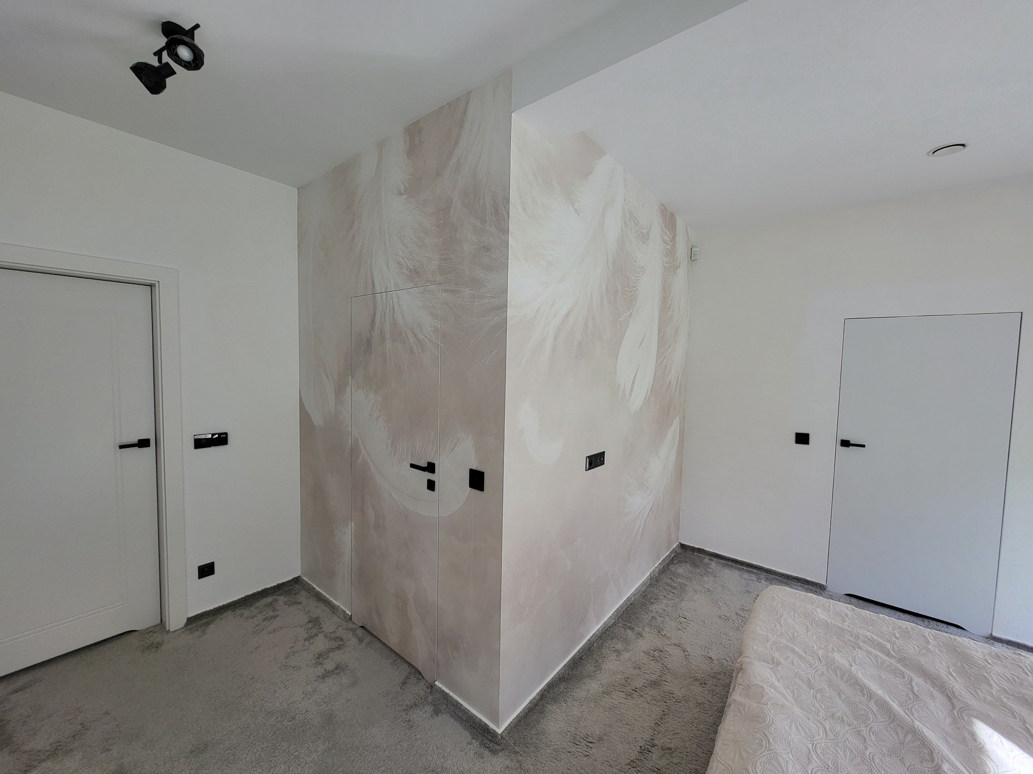 Pokój po remoncie: ściana z tapetą w pióra, minimalistyczne białe drzwi z czarnymi klamkami, szara wykładzina. Nowoczesne oświetlenie sufitowe.