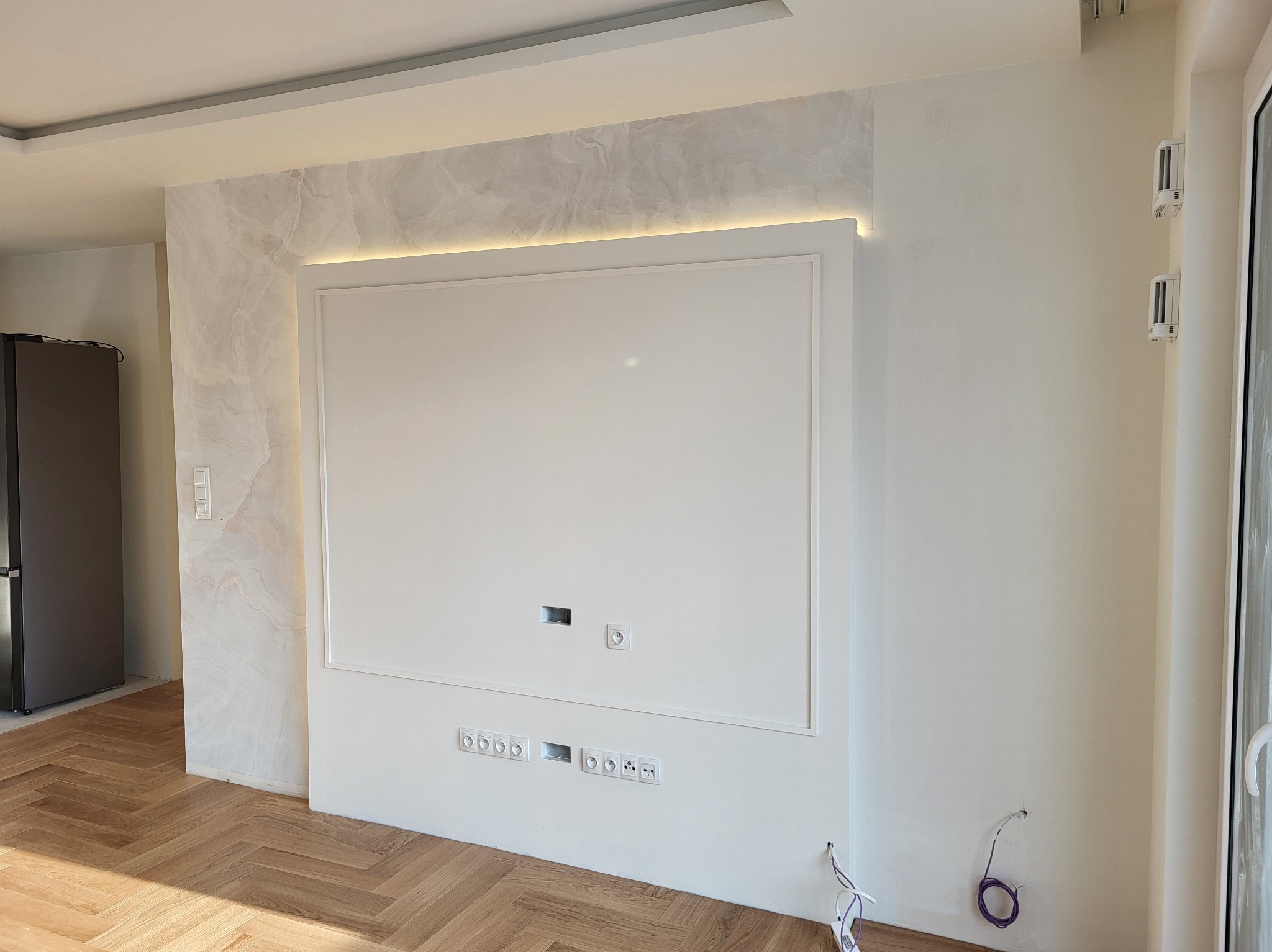 Wykończona ściana z dekoracyjnym panelem TV w stylu marmuru i białym obramowaniem, podświetlona listwą LED. Parkiet w jodełkę na podłodze, gniazdka elektryczne w ścianie.