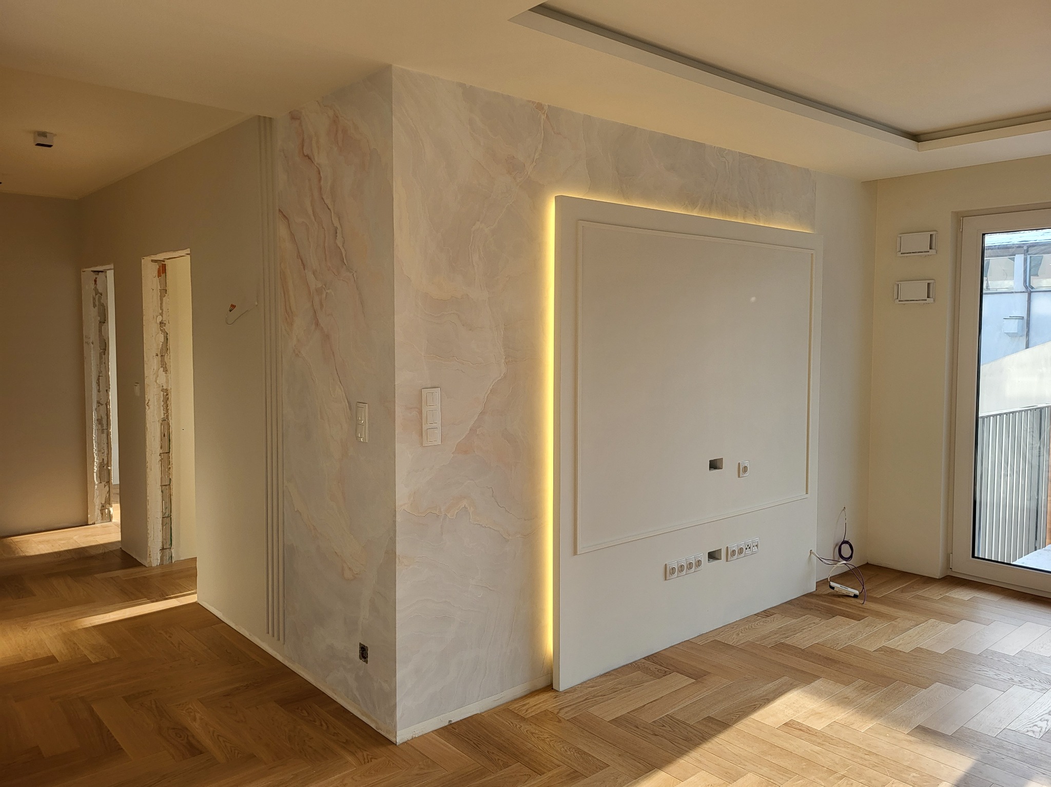 Wykończone wnętrze: parkiet w jodełkę, ściana z efektem marmuru z podświetleniem LED, biała ściana z gniazdkami i otwarta przestrzeń. Nowoczesny design.