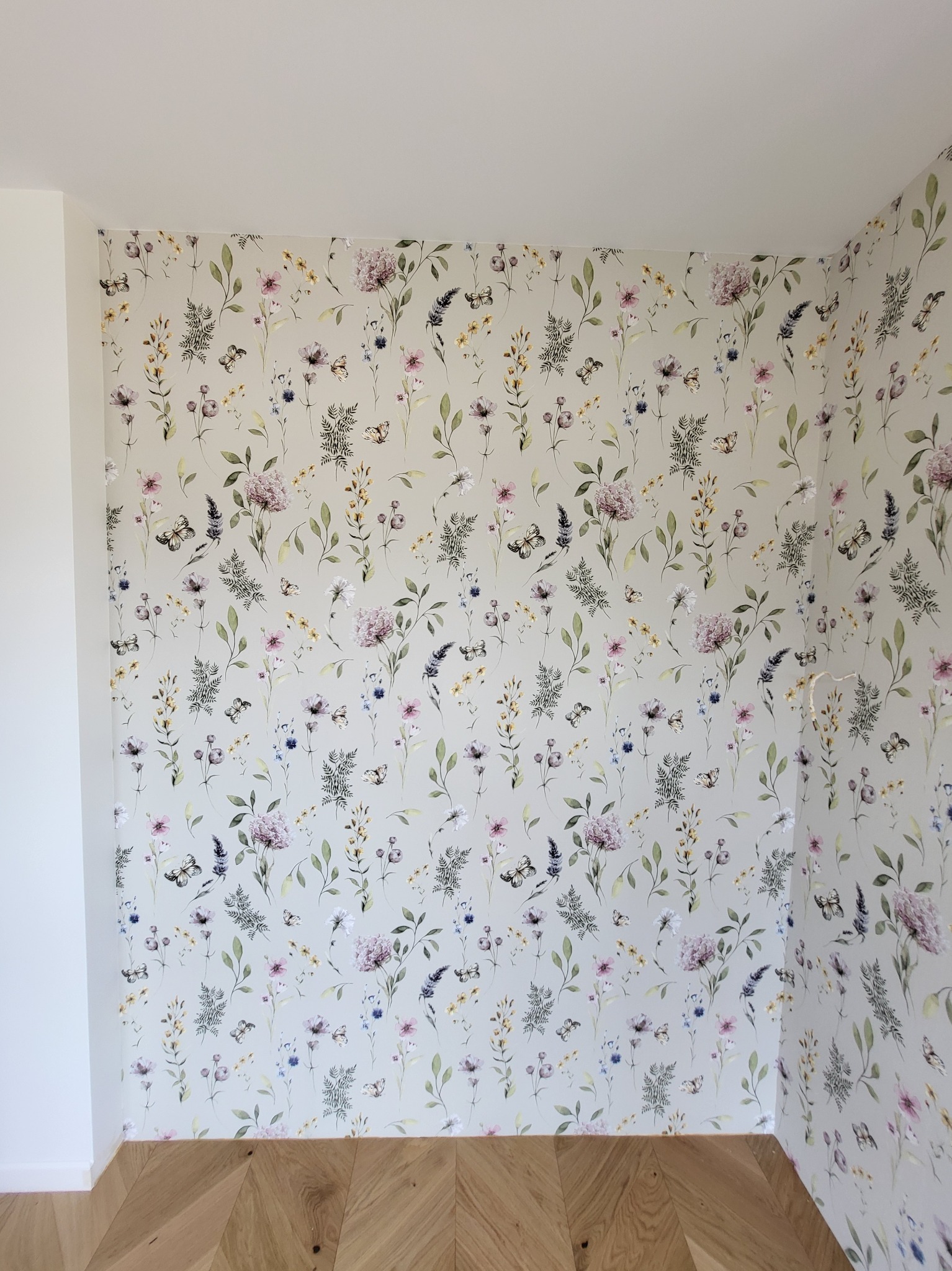 Ściana z tapetą w delikatne kwiaty i motyle, z widoczną podłogą z paneli ułożonych w jodełkę. Jasne, stonowane kolory i naturalne światło.