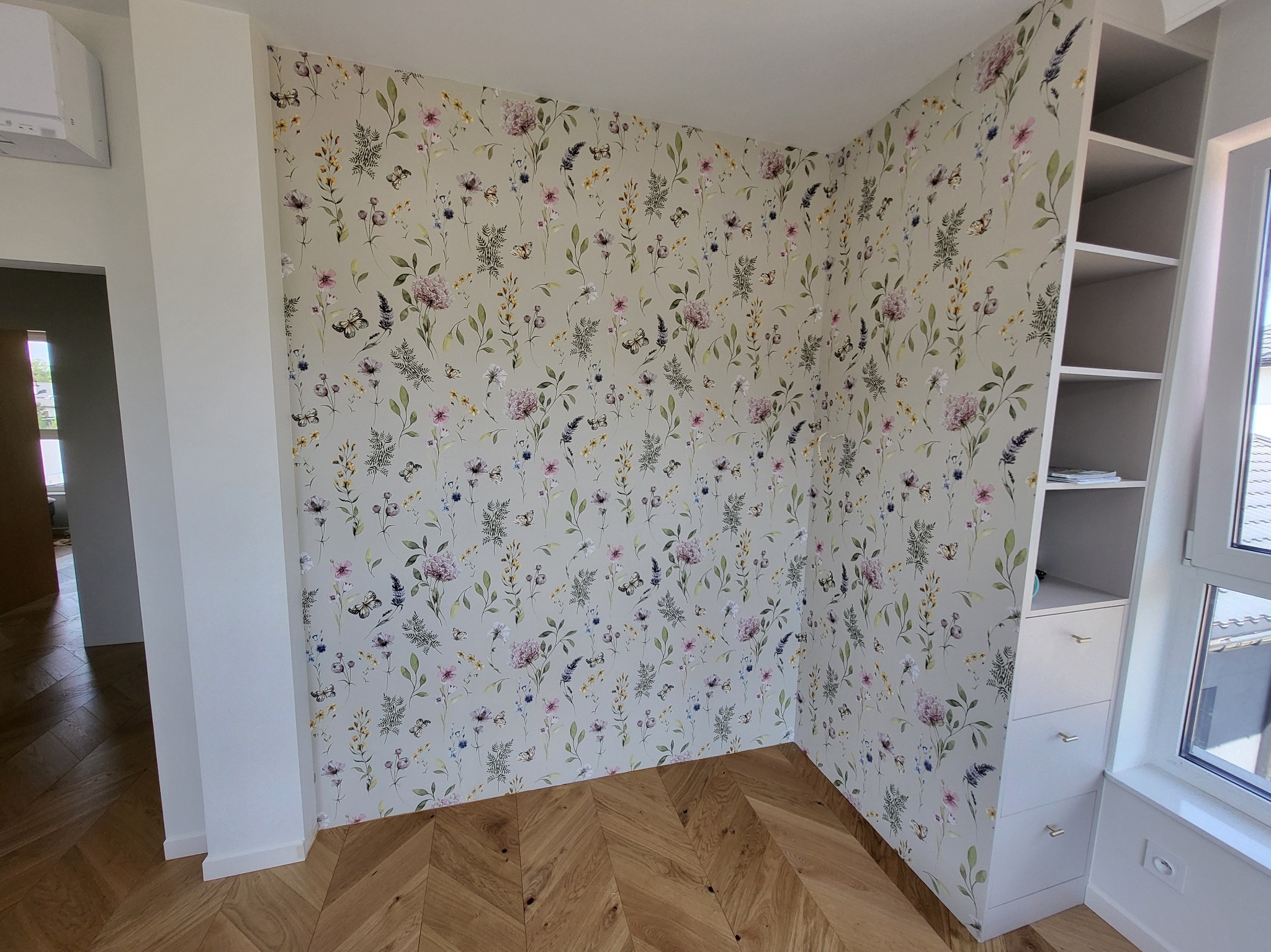 Pokój z tapetą w kwiaty i drewnianą podłogą w jodełkę. Przy oknie biały regał z szufladami. Neutralna kolorystyka, jasne wnętrze.
