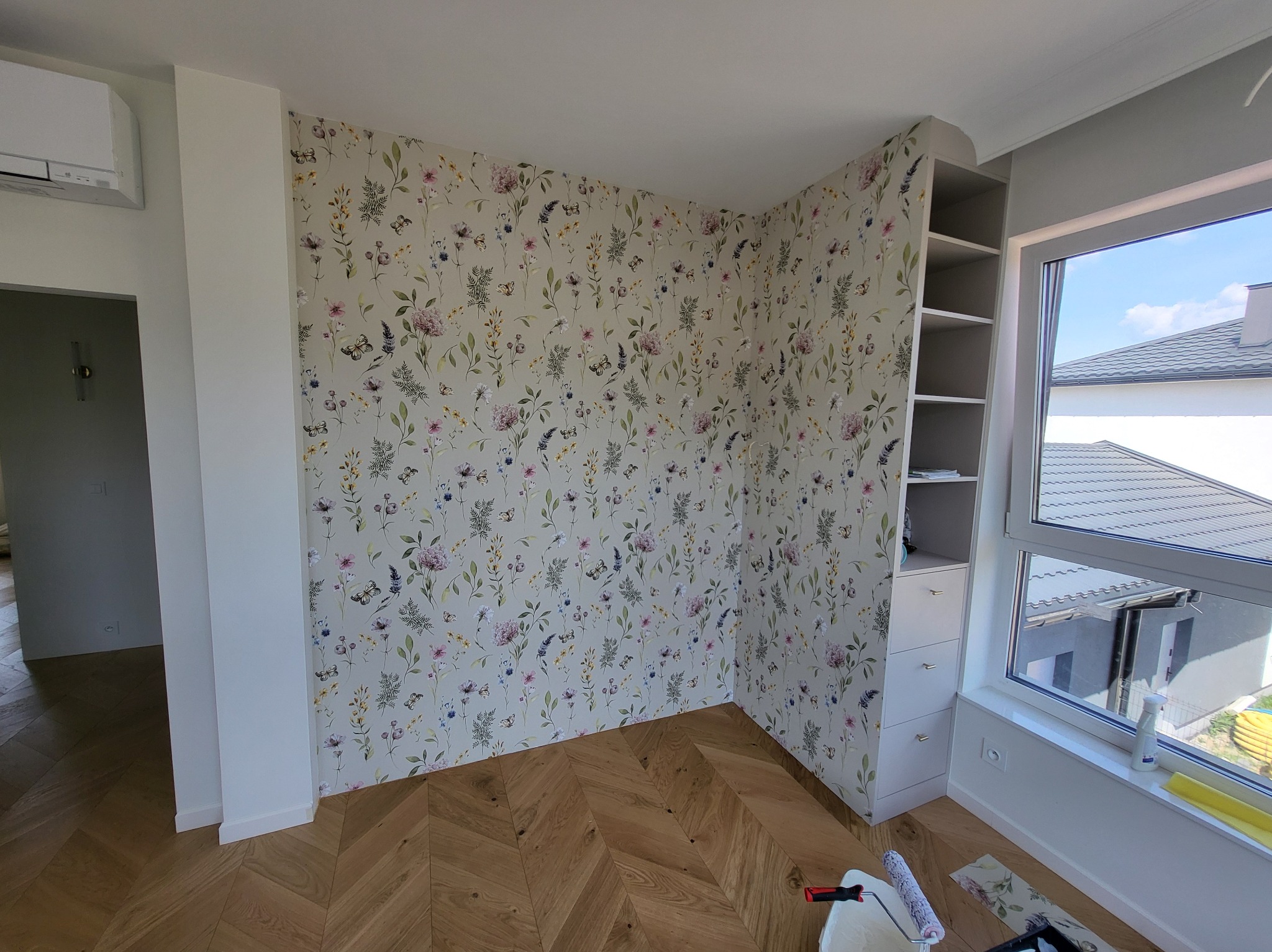 Pokój po tapetowaniu z motywem kwiatowym, parkiet w jodełkę, zabudowa meblowa przy oknie. Widoczne narzędzia do tapetowania.