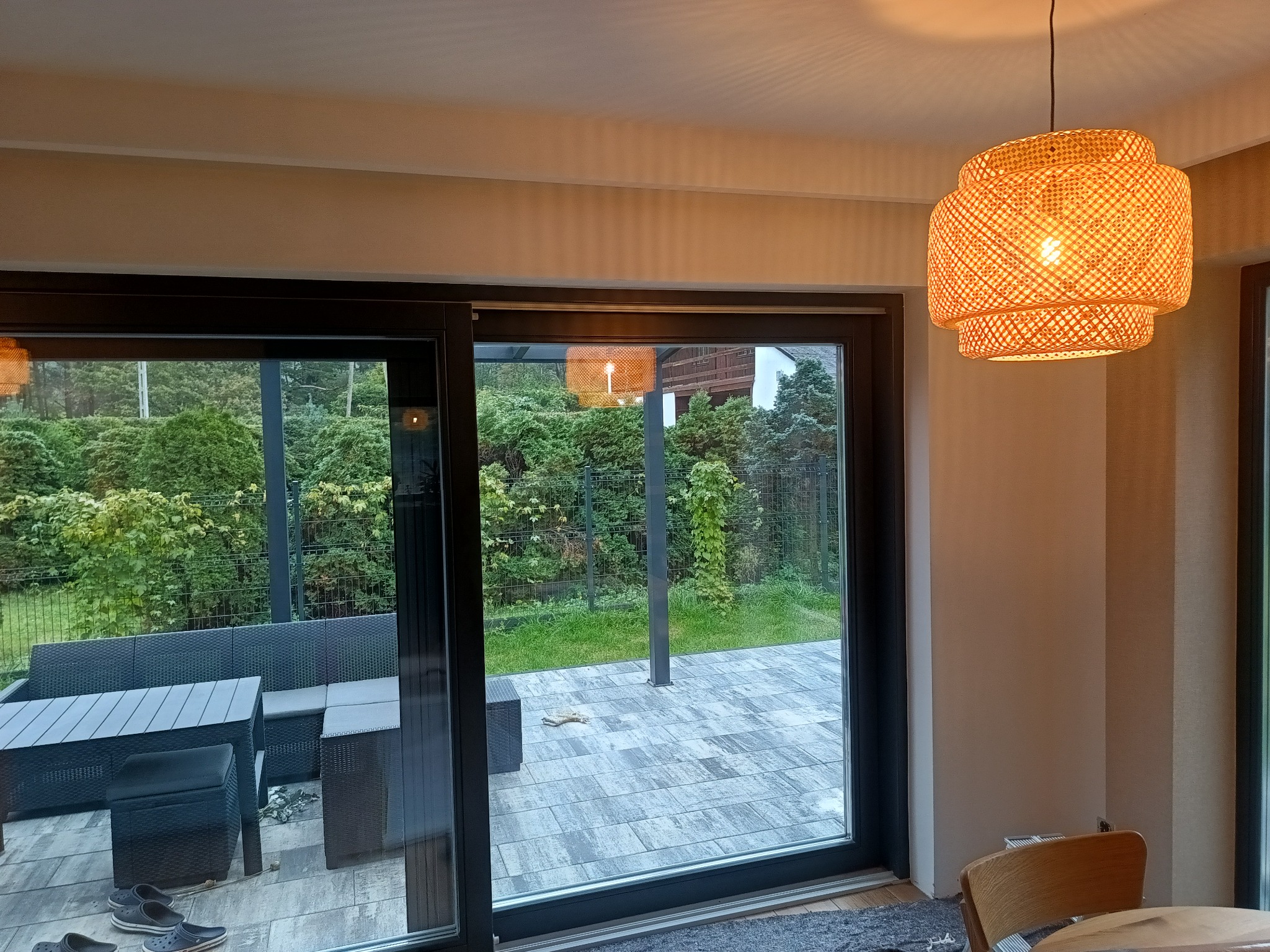 Jasne wnętrze z widokiem na taras przez przesuwne drzwi. W tle zielony ogród i rattanowa lampa wisząca. Minimalistyczny design.