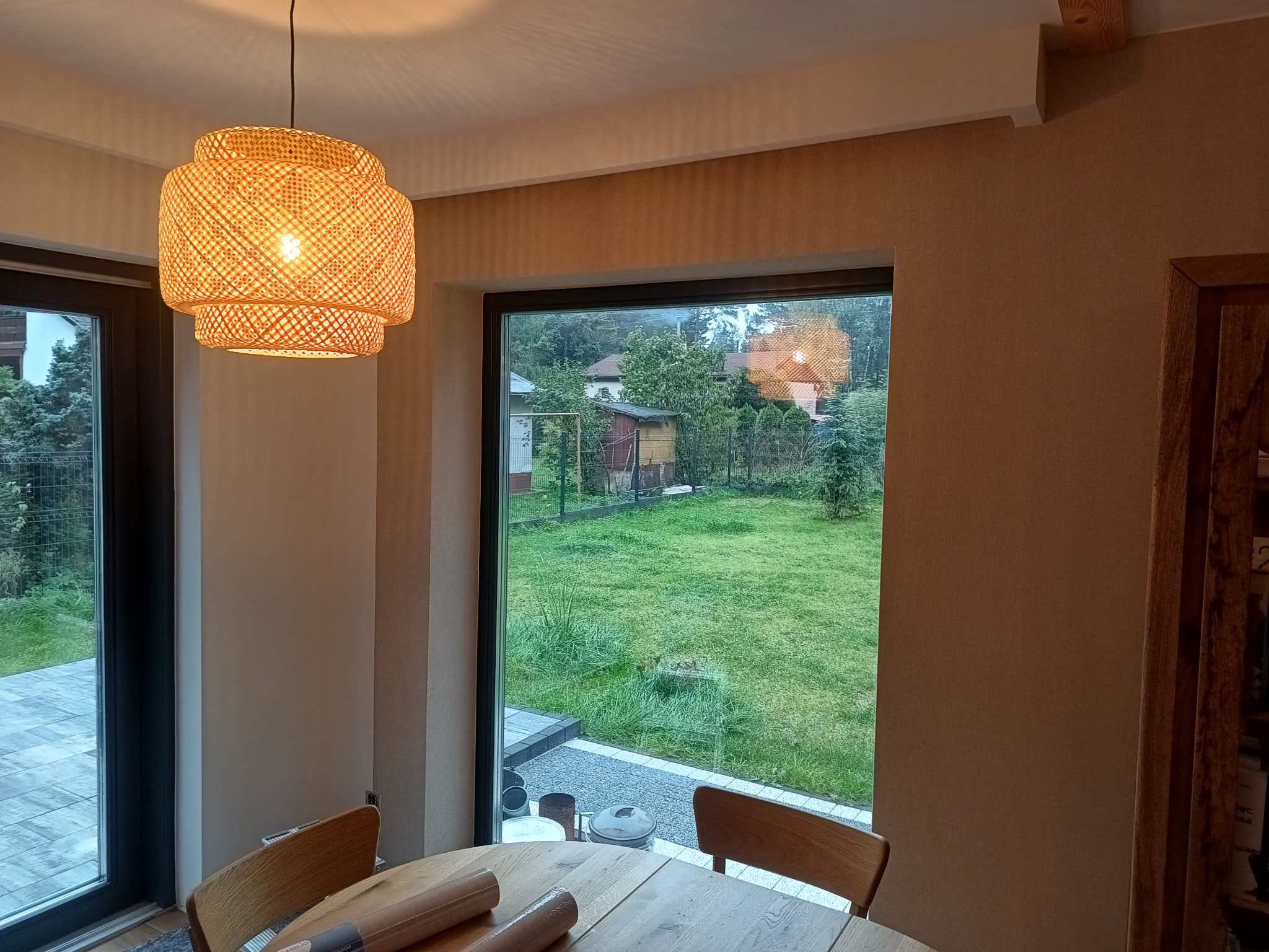 Jasne wnętrze z widokiem na ogród przez duże okno. Na pierwszym planie stół z rolkami tapety i pleciona lampa. Ściany wykończone tapetą o jednolitej fakturze.