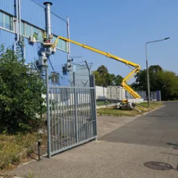 Pracownik w podnośniku koszowym przy niebieskiej hali przemysłowej, wykonujący prace na wysokości. Widoczna brama wjazdowa i fragment chodnika.