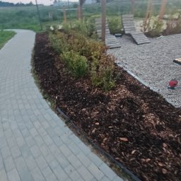 PPHU ARTEK ARTUR NOWAKOWSKI - Ścieżka z kostki brukowej obok rabaty z korą i krzewami, w tle drewniana konstrukcja placu zabaw, wieczorny krajobraz.