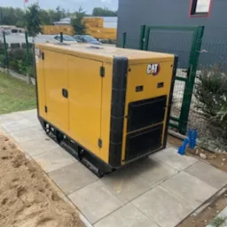 Żółty agregat prądotwórczy CAT ustawiony na betonowych płytach chodnikowych obok budynku przemysłowego i ogrodzenia z siatki.