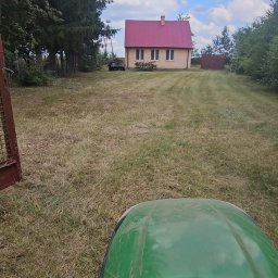 PPHU ARTEK ARTUR NOWAKOWSKI - Zielony traktor kosi trawę na posesji z domem o czerwonym dachu w tle. Widok z perspektywy operatora, brama z lewej strony kadru.