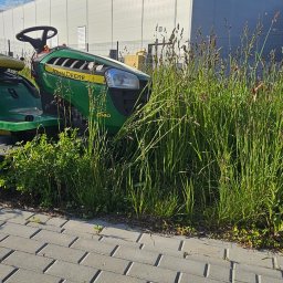 PPHU ARTEK ARTUR NOWAKOWSKI - Zielony traktor kosiarka John Deere D140 częściowo schowany w wysokiej trawie, na tle kostki brukowej i szarego budynku. Błękitne niebo z chmurami w tle.