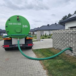 PPHU ARTEK ARTUR NOWAKOWSKI - Zielony wóz asenizacyjny z numerem telefonu podłączony wężem do otworu w fundamencie domu jednorodzinnego na tle zabudowy osiedlowej.