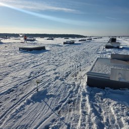 PPHU ARTEK ARTUR NOWAKOWSKI - Pracownik w kamizelce odblaskowej odśnieża płaski dach pokryty śniegiem, używając czerwonej łopaty. Widoczne metalowe elementy konstrukcyjne i ślady odśnieżania.
