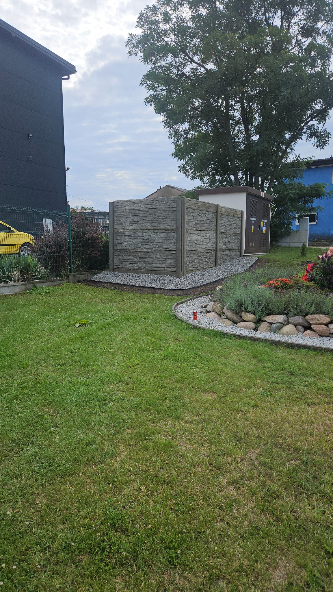 Nowoczesny, panelowy płot betonowy na posesji, otoczony szarym żwirem i zielenią trawnika, z fragmentem budynku i drzewa w tle. Estetyczne wykończenie ogrodu.