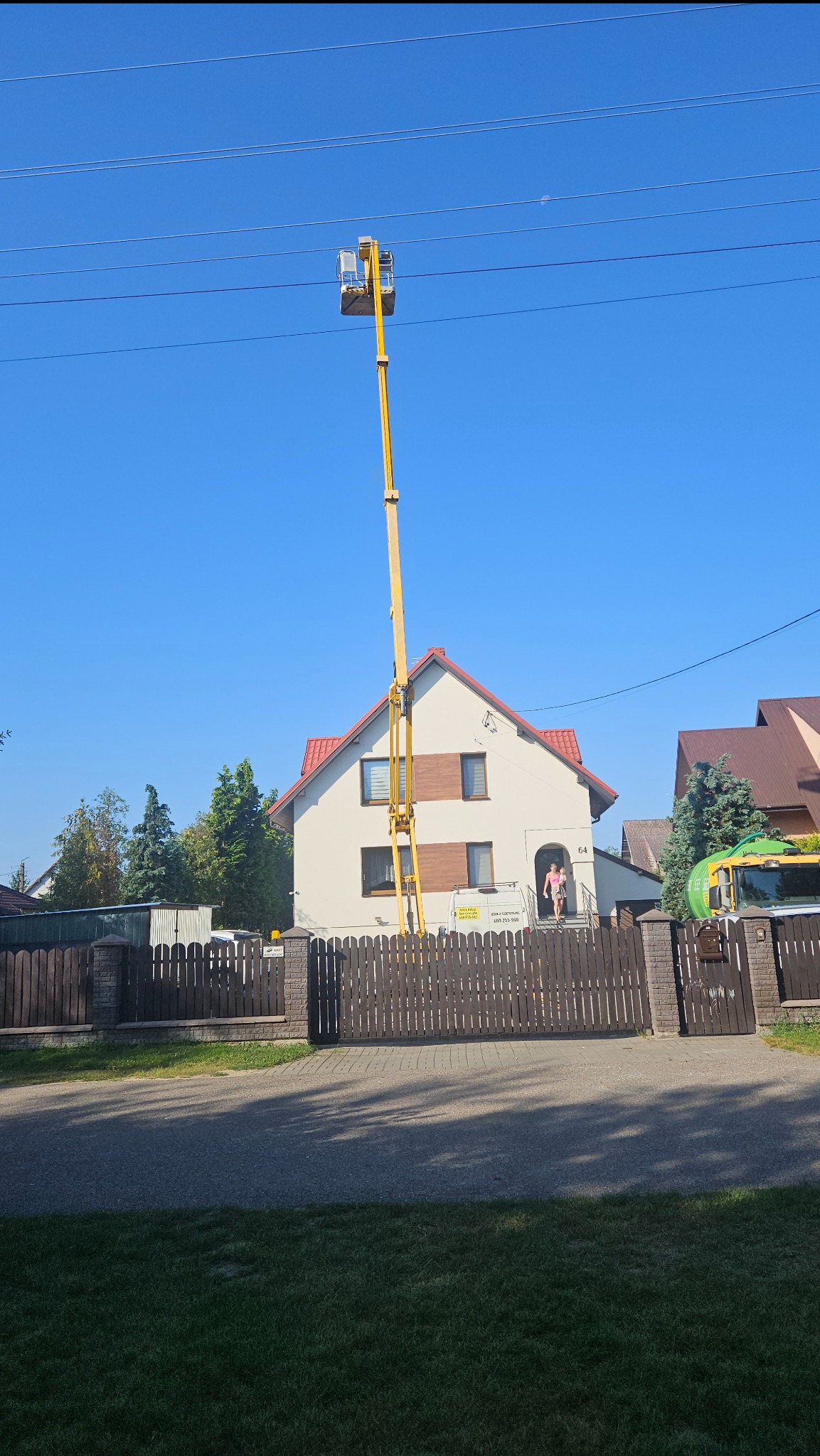 Żółty podnośnik koszowy przed domem z czerwoną dachówką, widoczne linie energetyczne i osoba w drzwiach. Błękitne niebo w tle.