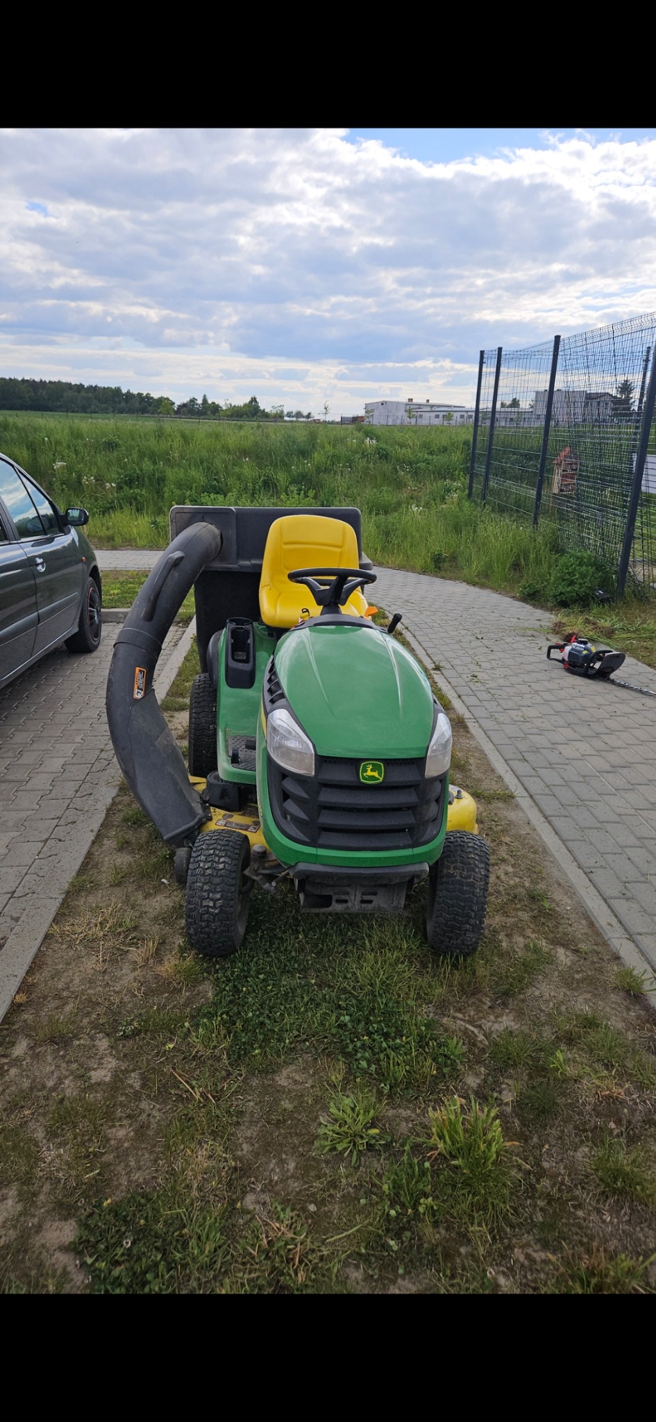 Traktorek do koszenia trawy John Deere z koszem, zaparkowany na trawniku obok chodnika i ogrodzenia. W tle widoczne pole i budynki.