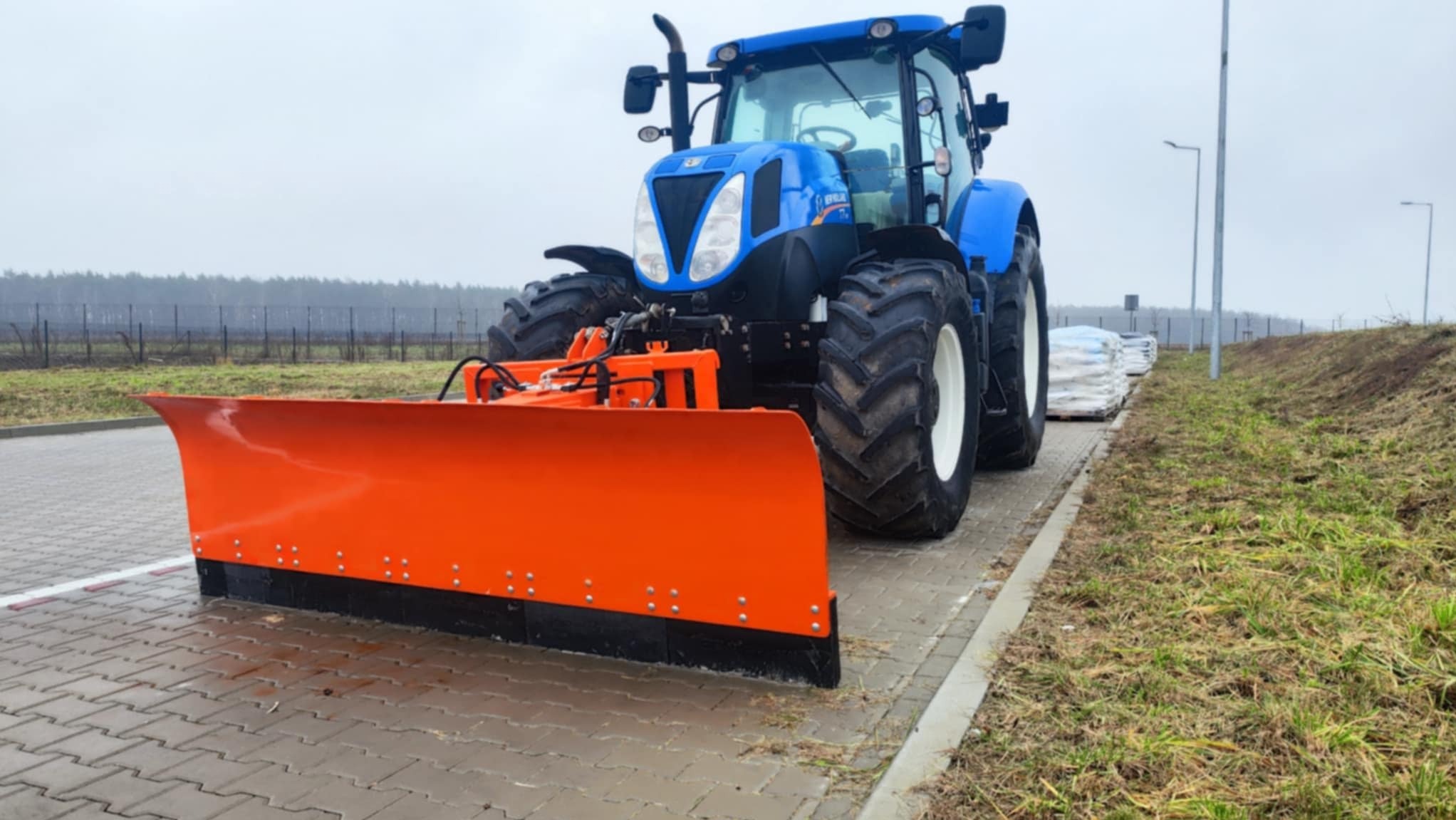 Niebieski traktor z pomarańczowym pługiem na kostce brukowej, worki w tle. Sprzęt do niwelacji terenu lub odśnieżania. Pochmurny dzień.