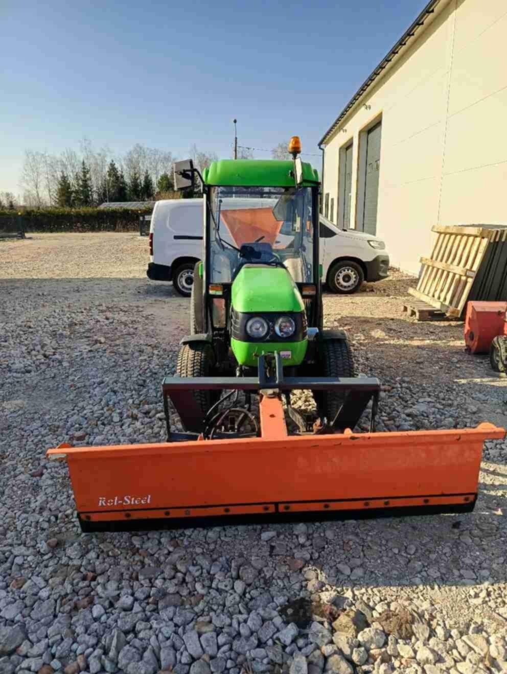 Zielony traktor z pomarańczowym pługiem Rel-Steel na żwirowym terenie, w tle budynek i białe busy. Sprzęt gotowy do pracy.