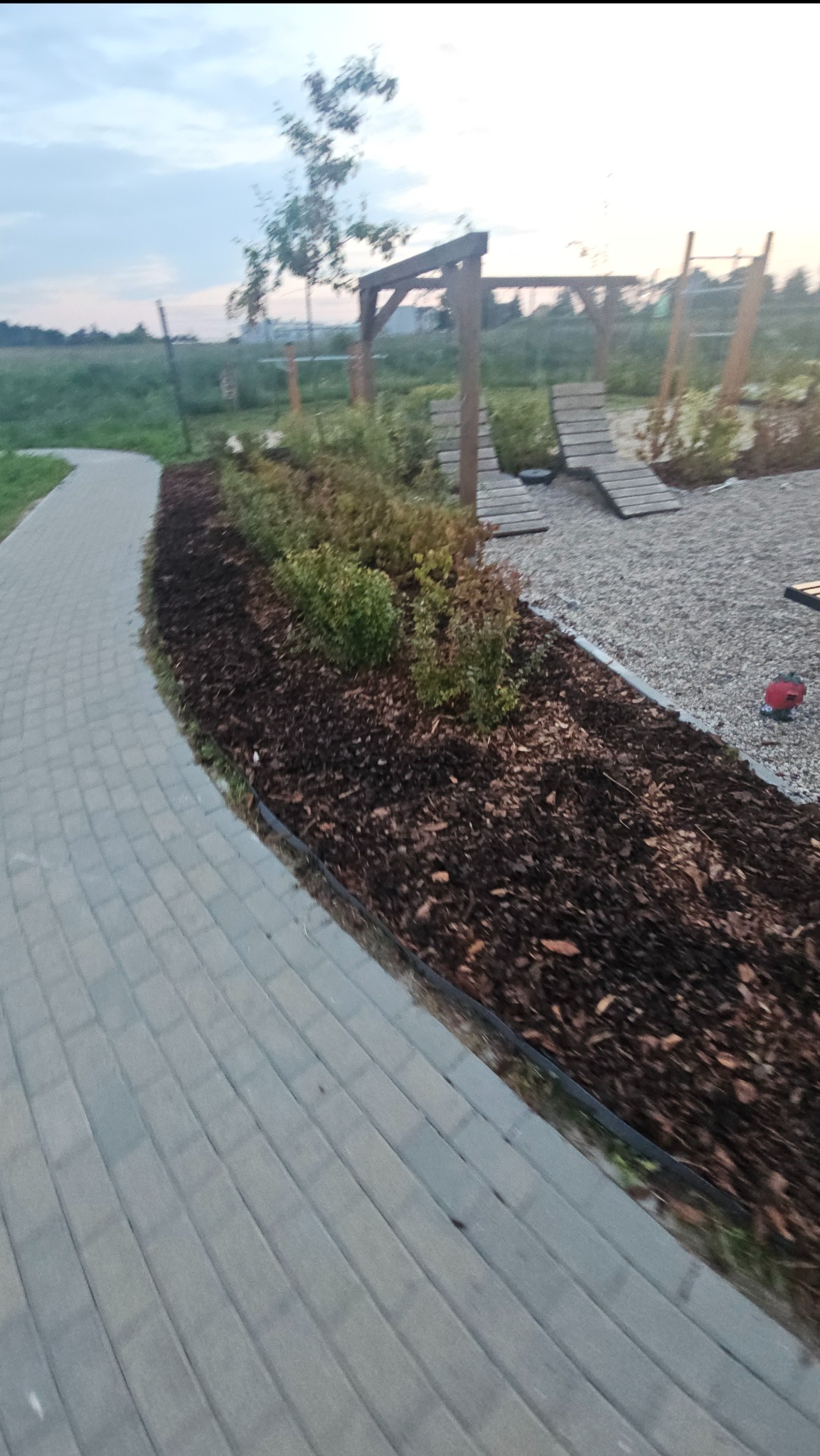 Ścieżka z kostki brukowej obok rabaty z korą i krzewami, w tle drewniana konstrukcja placu zabaw, wieczorny krajobraz.