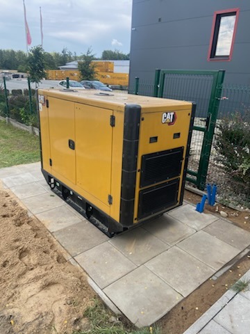 Żółty agregat prądotwórczy CAT ustawiony na betonowych płytach chodnikowych obok budynku przemysłowego i ogrodzenia z siatki.