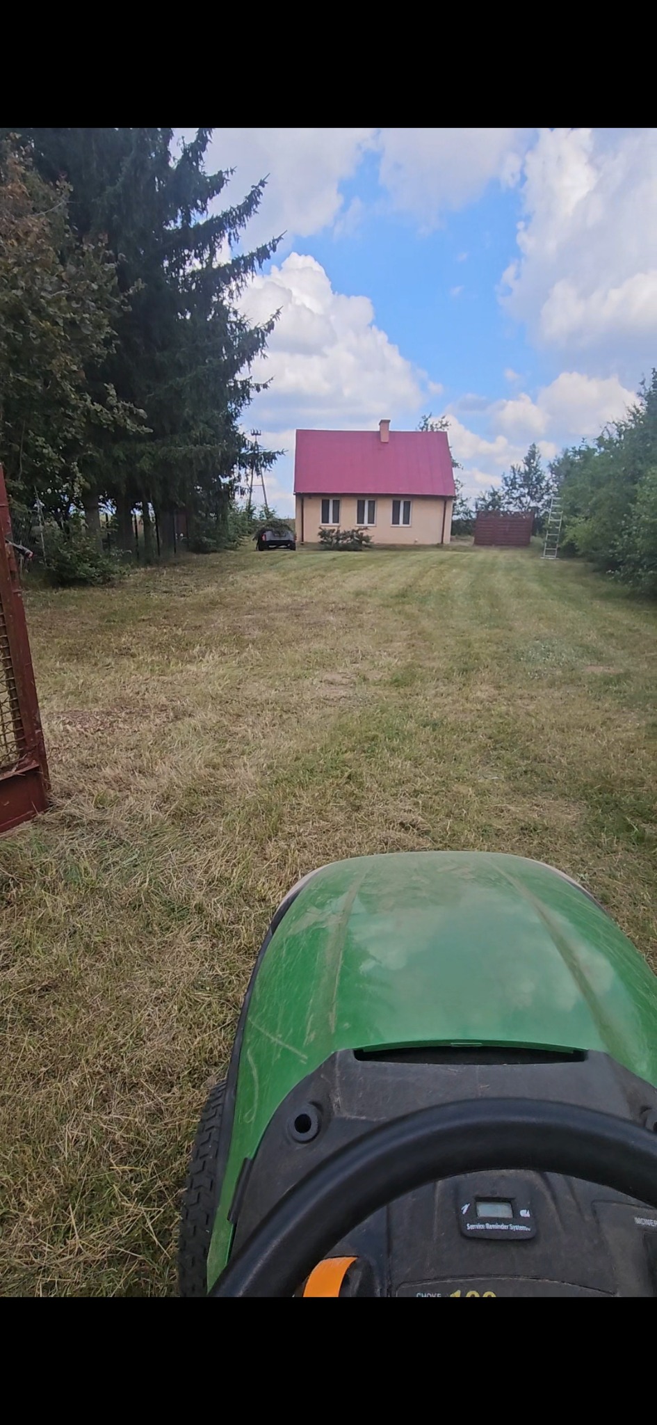 Zielony traktor kosi trawę na posesji z domem o czerwonym dachu w tle. Widok z perspektywy operatora, brama z lewej strony kadru.