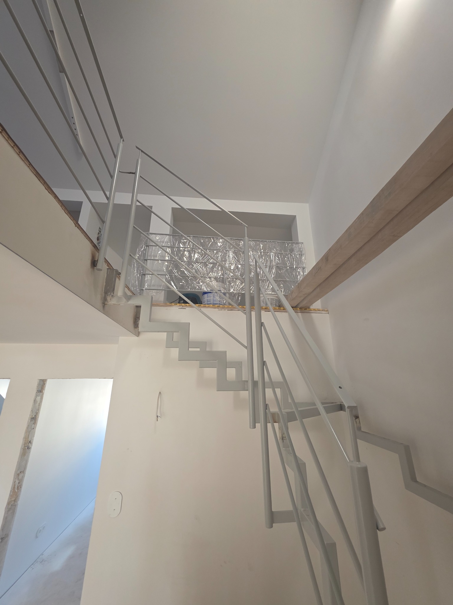 Nowoczesne, metalowe schody z minimalistyczną balustradą w jasnym wnętrzu. Schody pomalowane na szaro, zabezpieczone folią na górnym podeście. Widok z dołu.